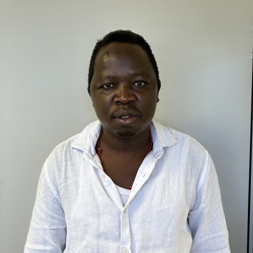 Person wearing a white button-up shirt with a visible necklace, seated against a plain light-colored background.