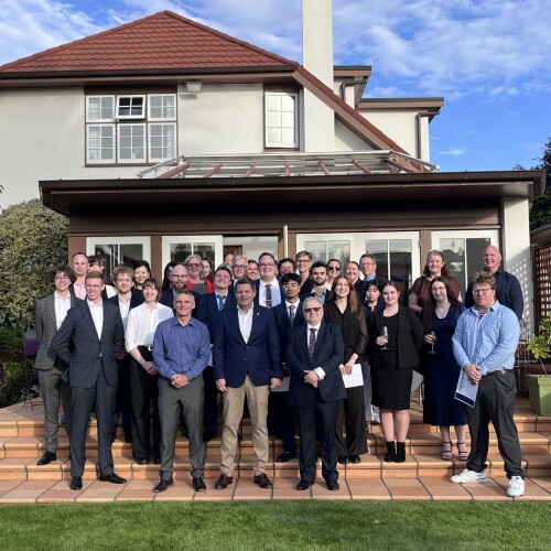 A group photo taken outdoors in front of a large house with a red-tiled roof and cream walls. The group consists of approximately 25 people standing on terracotta steps, dressed in formal and semi-formal attire. Some individuals are holding papers or drinks, and the setting appears to be a garden with green grass and clear blue sky in the background.