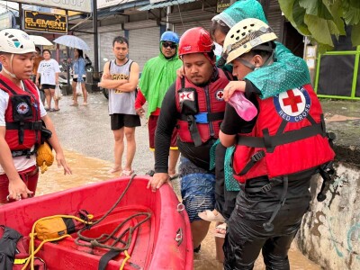 Red Cross rescue workers wearing helmets and life vests assist during a flood response, standing next to a red inflatable rescue boat with ropes and equipment. Floodwater covers the street, and several people in rain gear and casual clothing are visible in the background near closed shopfronts.