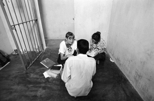 Two women sitting across from a man in a concrete cell.