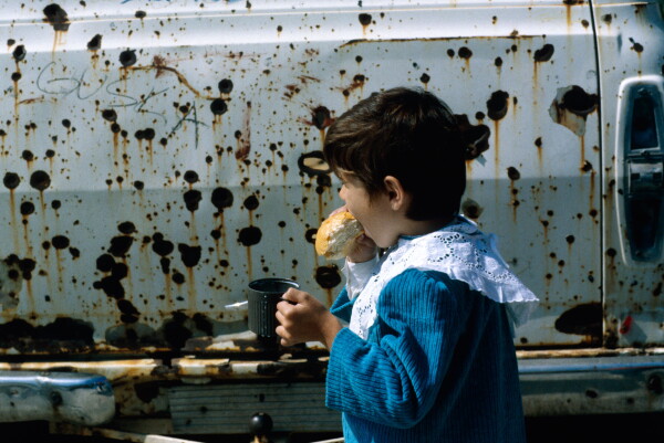 A child eating bread in front of a vehicle covered in bullet holes.