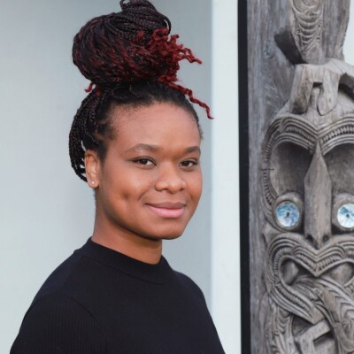 Person wearing a black long-sleeve top with intricately braided hair styled in a high bun featuring red highlights, standing next to a carved wooden figure with detailed patterns and inlaid blue circular elements. The background includes a light-colored wall and part of a dark frame.