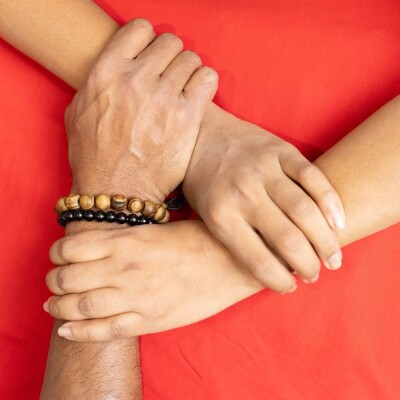 Two hands resting on a bright red surface, with one hand wearing two beaded bracelets—one in natural wood tones and the other in black—while the other hand gently holds the wrist.
