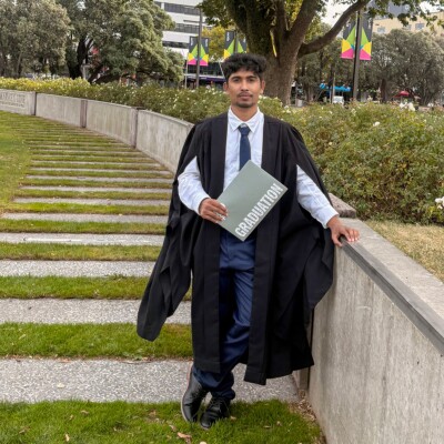 Person dressed in graduation attire including a black gown and tie, holding a folder labeled “GRADUATION,” standing on a pathway in a landscaped park with trees, grass, and buildings in the background.