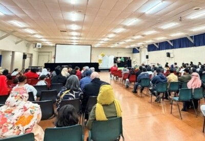 A large community gathering seated in rows of chairs inside a hall, facing a stage and projection screen during a World Refugee Day event in Palmerston North.