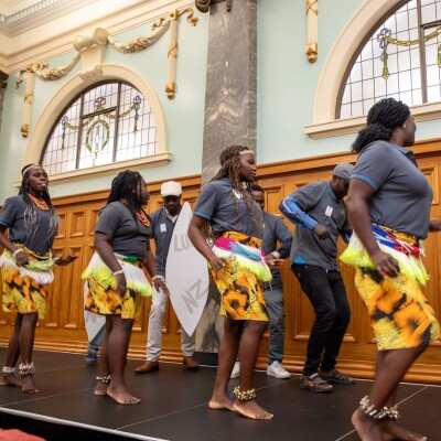 Performers take part in a cultural dance presentation on stage, wearing coordinated costumes and moving in formation against a backdrop of decorative windows and wood-panelled walls.