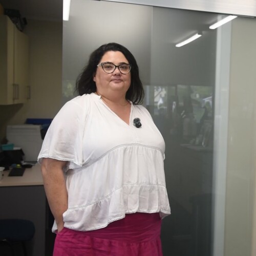 Person standing indoors near a glass partition, wearing a white top and pink skirt.
