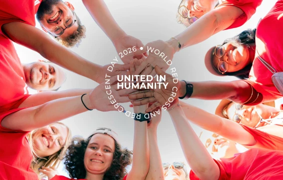 Group of Red Cross Red Crescent volunteers in red shirts forming a circle and stacking their hands together at the centre, viewed from below against a bright sky, with the text “United in Humanity – World Red Cross and Red Crescent Day 2026.
