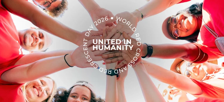 Group of Red Cross Red Crescent volunteers in red shirts forming a circle and stacking their hands together at the centre, viewed from below against a bright sky, with the text “United in Humanity – World Red Cross and Red Crescent Day 2026.