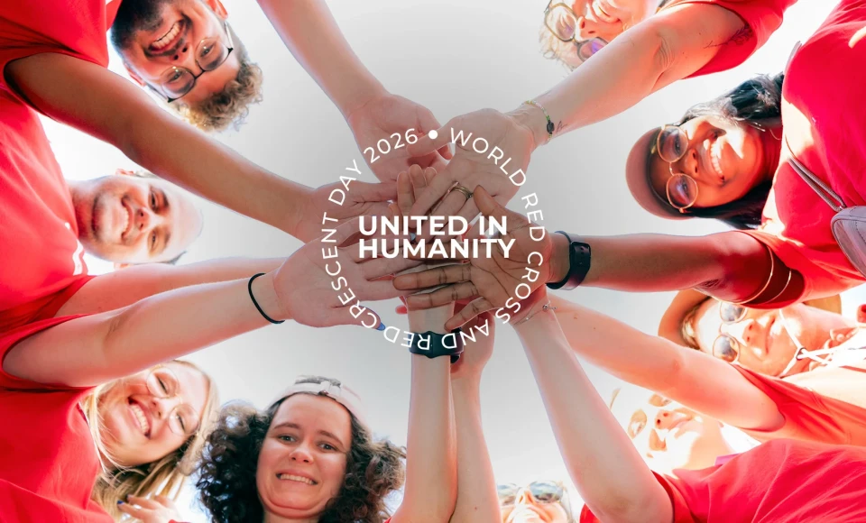 Group of Red Cross Red Crescent volunteers in red shirts forming a circle and stacking their hands together at the centre, viewed from below against a bright sky, with the text “United in Humanity – World Red Cross and Red Crescent Day 2026.
